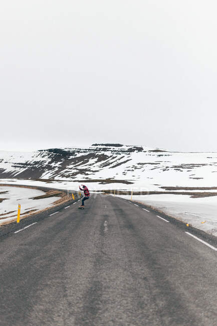 Rückansicht des Hipster-Typs Skateboard fahren auf gepflasterter langer Straße mit schneebedeckten Bergen im Hintergrund in Island. — Stockfoto