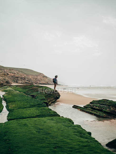 Человек стоит на зеленом камне у океана — стоковое фото