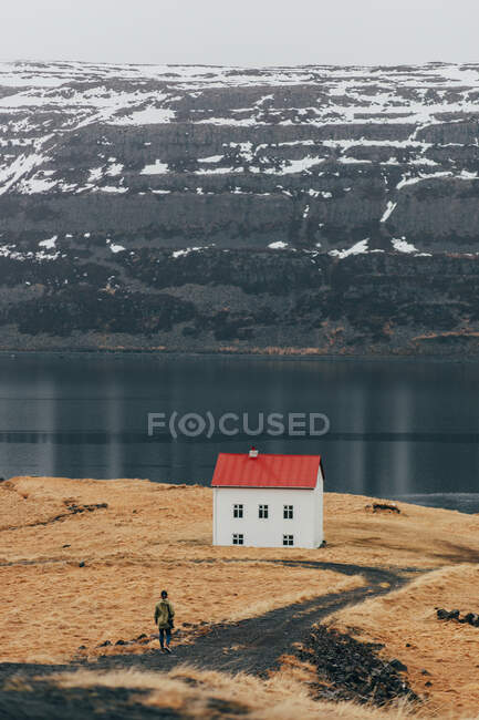 View of tourist walking on road towards small house placed alone on coast of cold lake in Iceland. — Stock Photo
