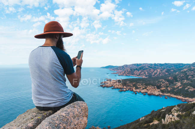 Hombre con sombrero sentado en la roca a la orilla del mar y tomando fotos - foto de stock