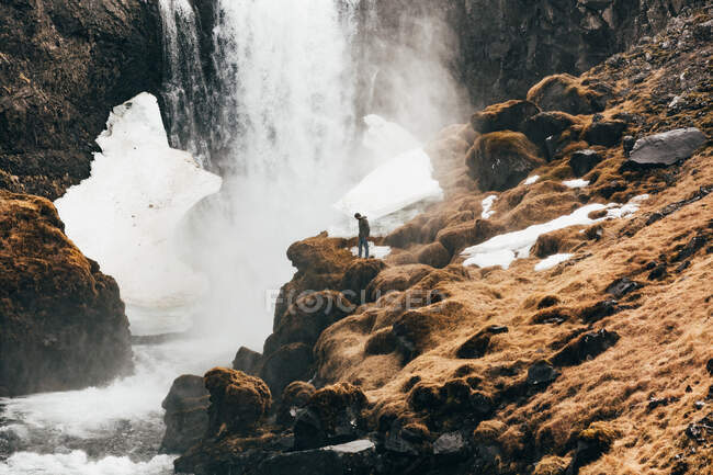 Vista a distancia del hombre de pie en el borde rocoso de la colina con salpicaduras de cascada en el fondo, Islandia. - foto de stock