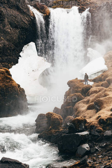 Вид на человека, стоящего на скалистом краю холма, на фоне которого плещется водопад, Исландия. — стоковое фото