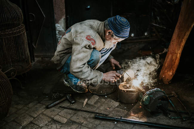Side view of man in dirty clothes welding items with sparks and smoke out sitting on ground. — Stock Photo