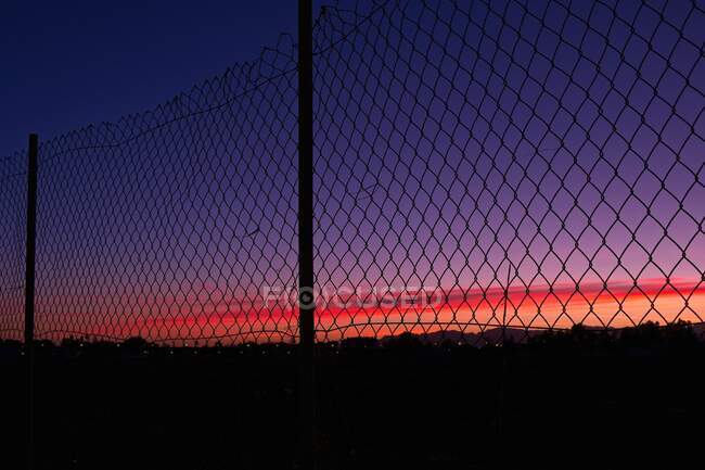 Silhueta de vista perto da cidade ao pôr do sol através de uma cerca — Fotografia de Stock