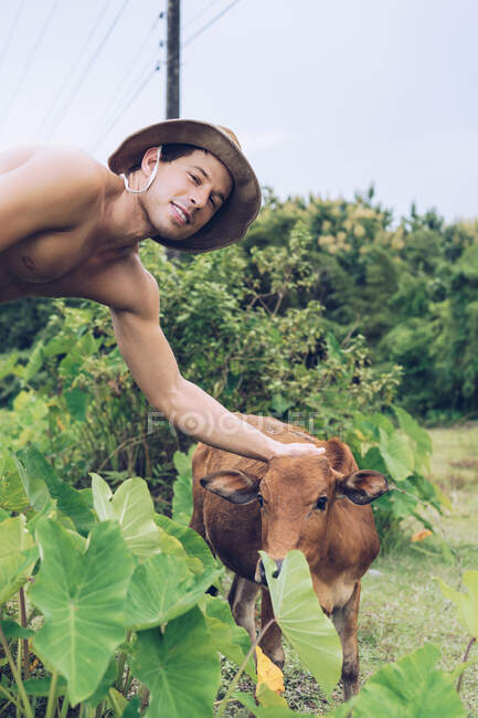 Adult shirtless tourist man stroking small brown calf in nature. — Stock Photo