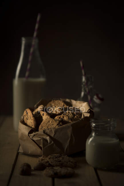 Cookies with chocolate in paper bundle and bottle of milk standing on wooden table on a dark background — Stock Photo