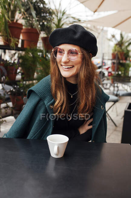 Ставтеся до молодої привабливої жінки в береті і сонцезахисних окулярах сидячи в кафе і озираючись. — стокове фото
