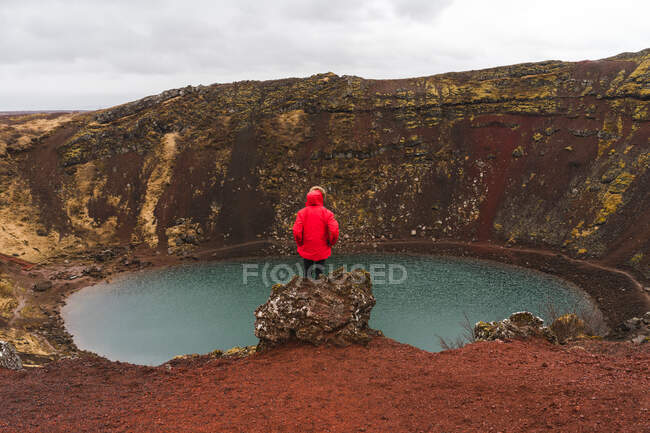 Back view of faceless man in warm clothes sitting on rim of open pit and looking at small lake while traveling through Iceland. - foto de stock
