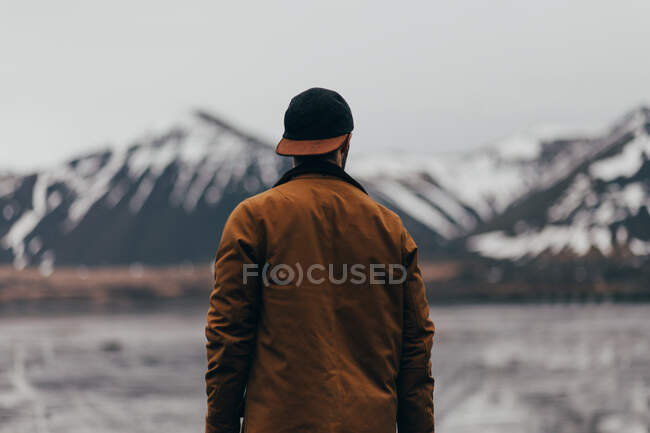 Back view of traveler in brown jacket and cap standing against cold mountains of Iceland. — Stock Photo