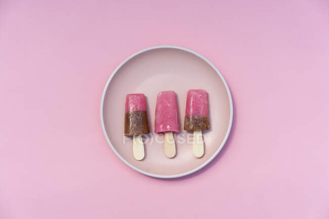 Colorful pink and brown popsicles lying in row on pastel plate on pink background from above — Stock Photo