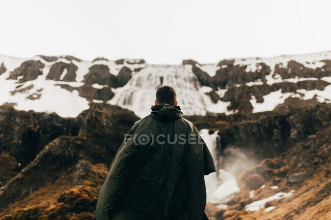 Rückenansicht eines Mannes im grünen Regenmantel, der auf einer Landschaft mit Bergen und Wasserfall in Island steht. — Stockfoto
