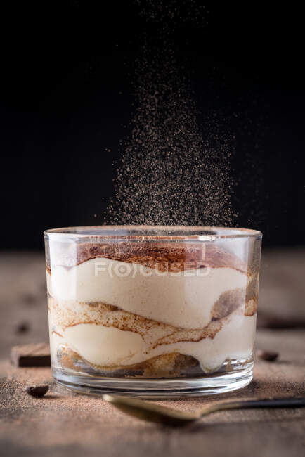 Primer plano del postre de tiramisú fresco en vidrio con partículas de polvo de cacao que caen encima. - foto de stock