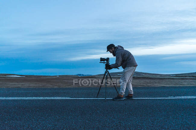 Vue latérale de l'homme en vêtements chauds mettant appareil photo professionnel sur trépied tout en prenant des photos de la nature islandaise. — Photo de stock