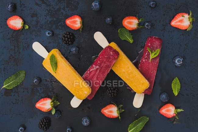 Vista de cima de sorvetes amarelos e vermelhos sobre fundo escuro com morangos amoras mirtilos e hortelã — Fotografia de Stock