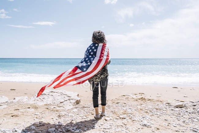Vista trasera de la joven mujer caminando hacia el mar con bandera de EE.UU. en la playa de arena . - foto de stock
