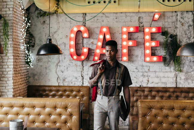Handsome african male in pants with suspenders and shirt putting hands in pockets standing and looking at camera — Stock Photo