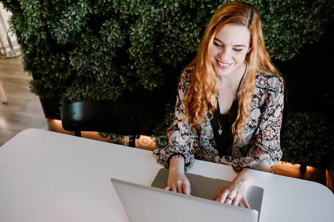 Joyeux jeune rousse femme assise à l'ordinateur portable et tapant à la table — Photo de stock