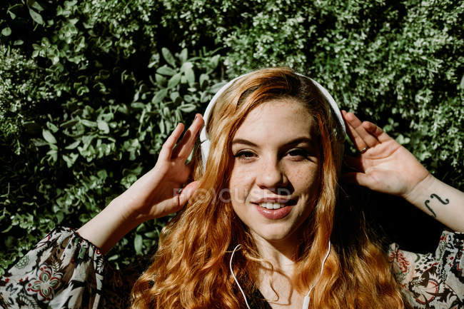 Lächelnde junge rothaarige Frau setzt Kopfhörer gegen Busch auf — Stockfoto
