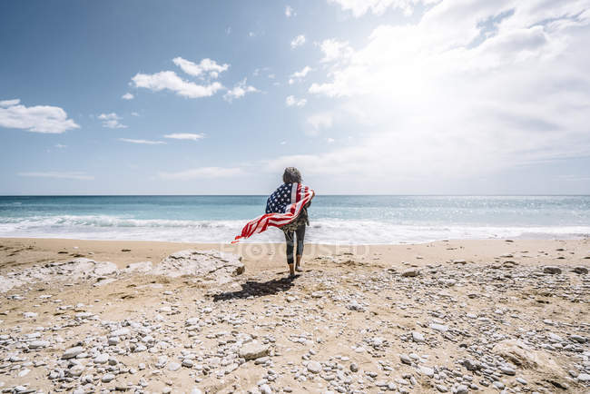 Vista posteriore della giovane donna che cammina verso il mare con bandiera USA sulla spiaggia sabbiosa . — Foto stock