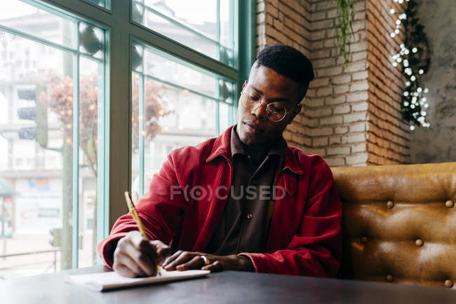 Hombre negro sentado en la cafetería y escribiendo - foto de stock