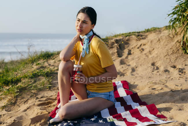 Дівчина з напоєм сидить на піску — стокове фото