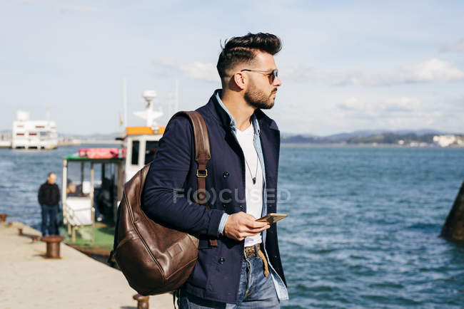 Человек с рюкзаком и смартфоном стоит в порту — стоковое фото