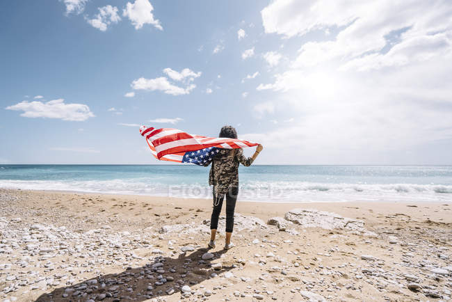 Rear view of young woman standing with USA flag on sandy beach. — Stock Photo