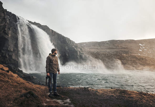 Seitenansicht eines Reisenden, der mit Fotokamera vor einem großen Wasserfall in Island steht und wegschaut. — Stockfoto