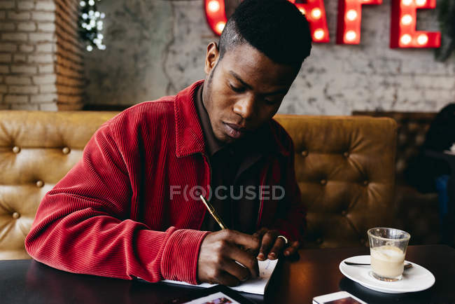 Hombre negro sentado en la cafetería y escribiendo - foto de stock
