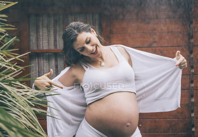 Smiling pregnant woman pointing on belly in rain against wooden house — Stock Photo