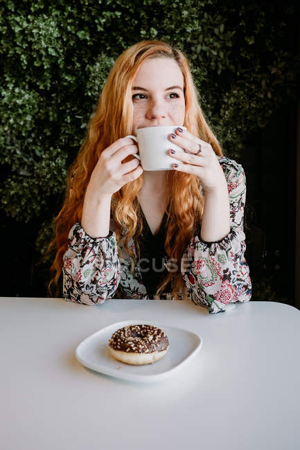 Усміхнена руда жінка з чашкою сидить і пончик сидить проти куща — стокове фото