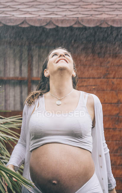 Heureuse femme enceinte debout sous la pluie contre la maison en bois — Photo de stock