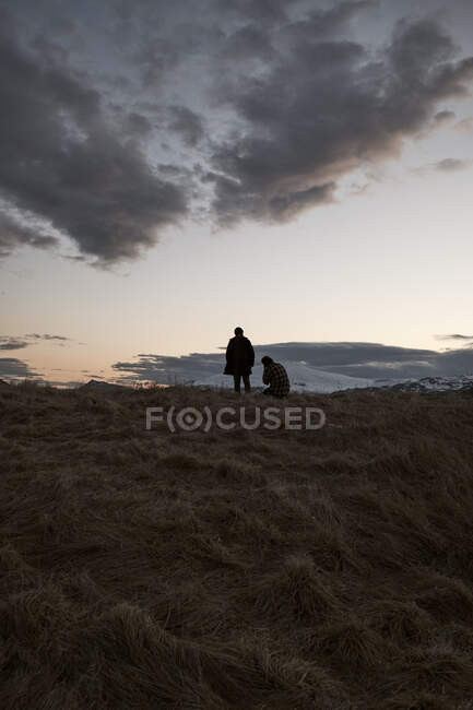 Deux voyageurs méconnaissables assis et debout sur de l'herbe sèche sous un ciel nuageux à flanc de colline en Islande — Photo de stock