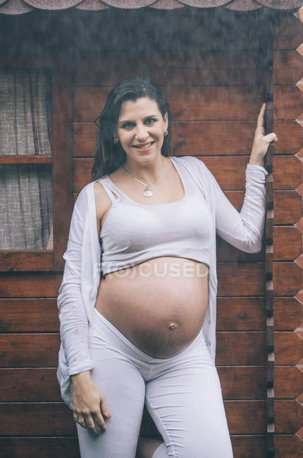 Усміхнена вагітна жінка стоїть під дощем проти дерев'яного будинку — стокове фото