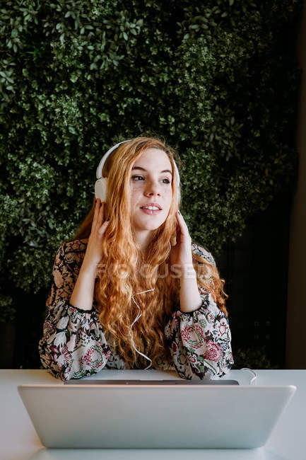 Jolie jeune rousse femme avec écouteurs assis avec ordinateur portable contre buisson — Photo de stock