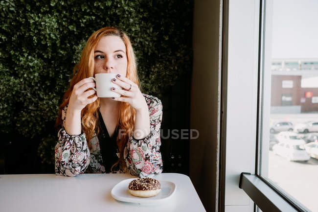 Улыбающаяся рыжая женщина с чашкой сидя и пончик сидя против куста — стоковое фото