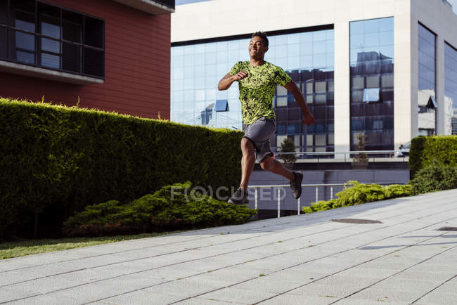 Cheerful sporty ethnic man in jump while running in city — Stock Photo