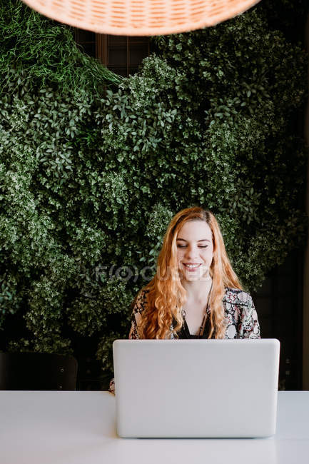 Sorridente bella rossa donna utilizzando il computer portatile a tavola contro cespuglio — Foto stock