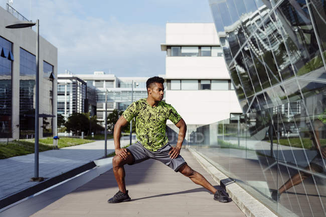 Hombre afroamericano calentando piernas en la ciudad con edificios modernos en el fondo - foto de stock