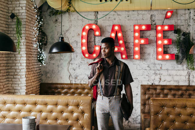 Handsome african male in pants with suspenders and shirt putting hands in pockets standing and looking at camera — Stock Photo