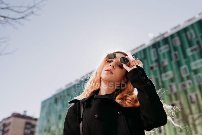 Стильная молодая рыжая женщина в солнцезащитных очках, стоящая в городе — стоковое фото
