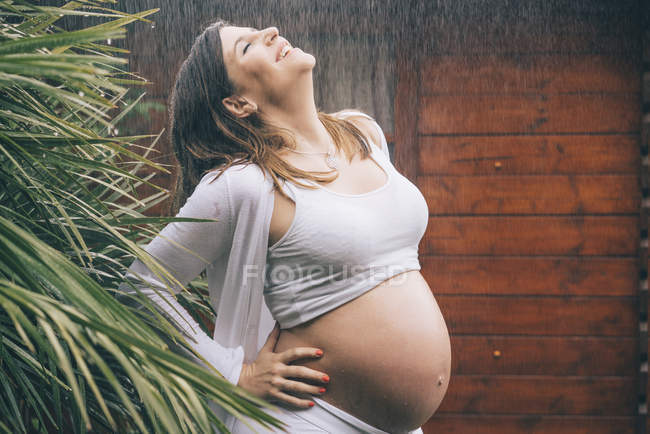 Happy pregnant woman standing in rain against wooden house — Stock Photo