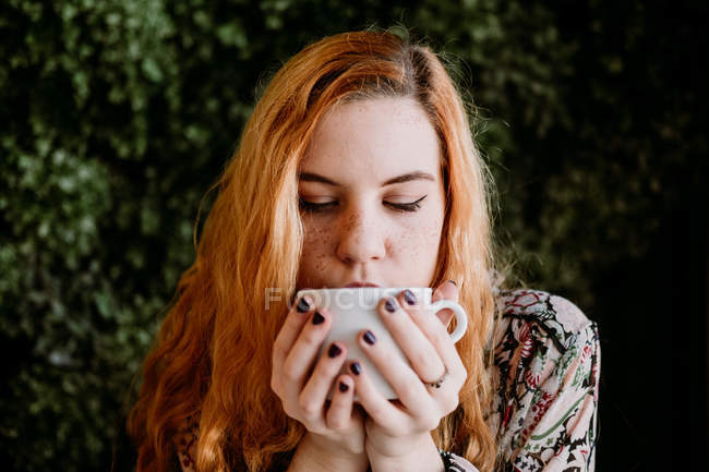 Рыжая молодая женщина пьет из чаши против куста — стоковое фото