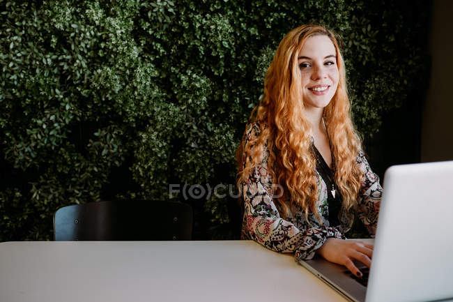Smiling pretty redhead woman using laptop at table against bush — Stock Photo