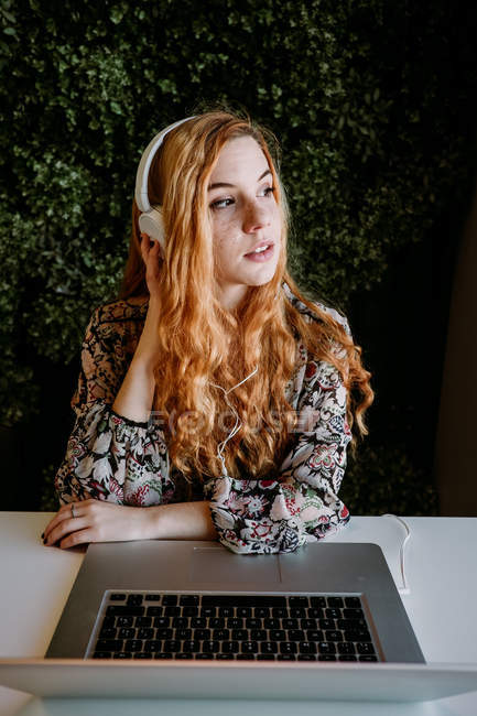 Jolie jeune rousse femme avec écouteurs assis avec ordinateur portable contre buisson — Photo de stock