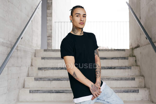 Joven hombre guapo con tatuajes en el cuello y las manos de pie en las escaleras mirando a la cámara - foto de stock