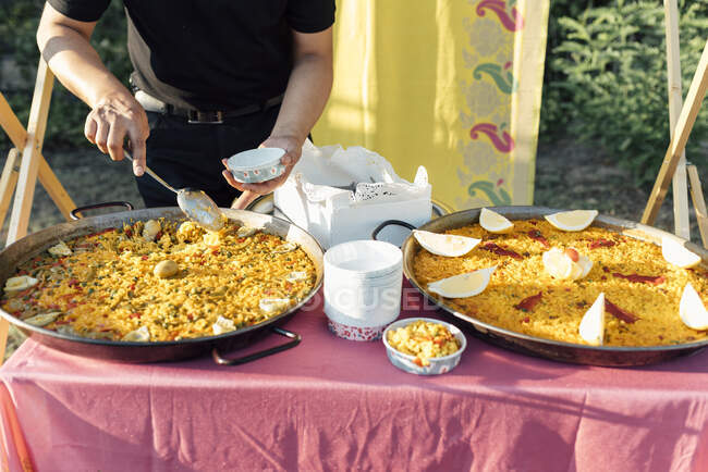 Crop unrecognizable cook standing at big pans and serving paella dish. — Stock Photo