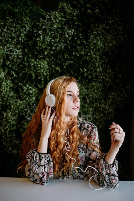 Jovem ruiva com fones de ouvido sentado à mesa contra arbusto — Fotografia de Stock