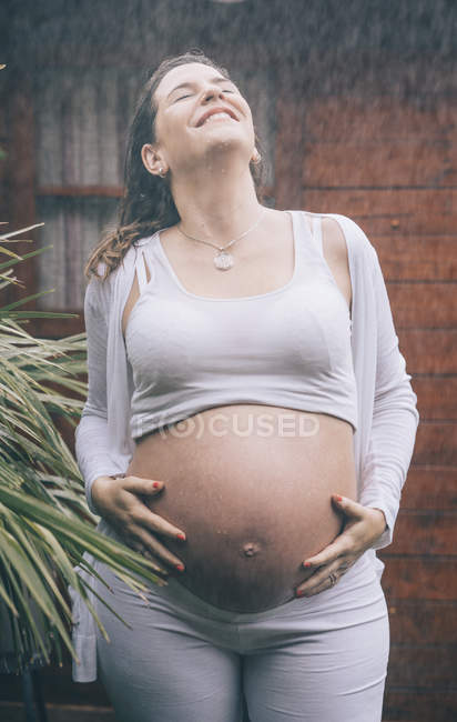 Mulher grávida feliz de pé na chuva contra a casa de madeira — Fotografia de Stock
