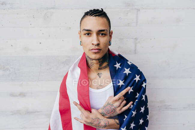 Латиноамериканське татуювання людини з пірсингом обгортаючи себе прапором об'єднаних станів і дивлячись на камеру на сірому текстурованому тлі — стокове фото